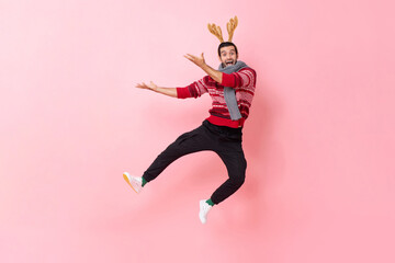 Full body festive portrait of Caucasian man wearing Christmas sweater and reindeer headband jumping...