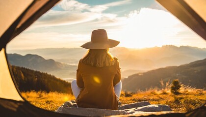 Camping in the wilderniss: Young woman with hat sitting in front of her tent on the moutain and enjoys the sunset in golden light. travel and adventure concept