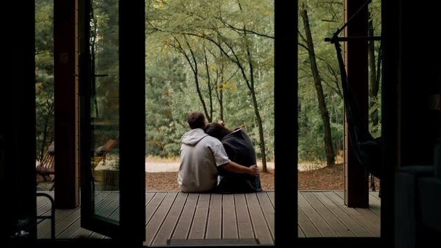 Man and woman in love sitting with their backs to the camera on a terrace overlooking  forest with plaids cuddling and talking