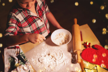 A little cute boy in red checkered pajamas is preparing Christmas gingerbread cookies on a festively decorated table.