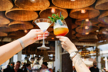 2 glasses of cocktails are cheers to life by female hands sitting at a beach bar on the boho light bulbs