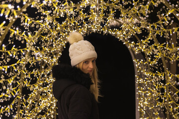 Portrait of beauty young woman in white hat and black jacket posing on tunnel made from decoration light. People outdoor photo