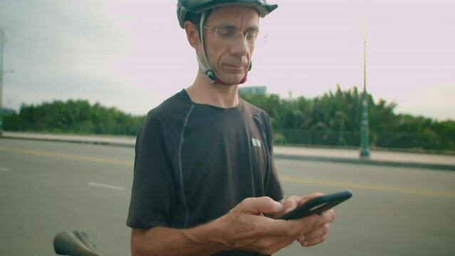 Medium shot of male cyclist in protective helmet and sportswear standing outdoors and using smartphone for navigation while taking break during bike ride