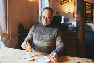 Indoor portrait of senior man drinking morning coffee with croissant