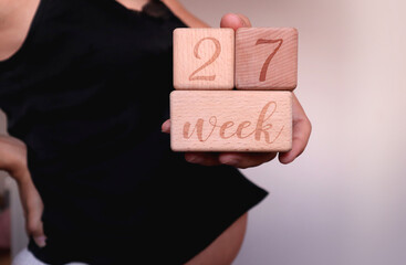 The pregnant woman in black top  touches her back and holds in hand wooden cubes with sign "27 weeks of pregnancy"