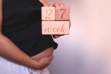 The pregnant woman in black top  touches her belly and holds in hand wooden cubes with sign "27 weeks of pregnancy"