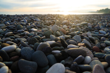 Pebble stones on the shore close up in the blurry sunset light in the distance background. sunrays. Pebble stones on sea shore. Blurred out of focus sun lights background, wallpaper. sun rays.