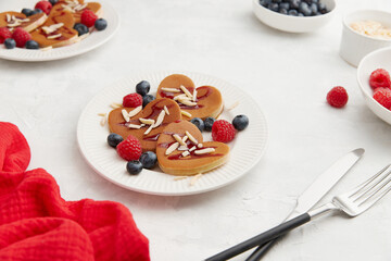 A heap of heart shaped pancakes with berries and cutlery on the white plate on grey background