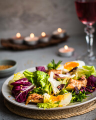 A bowl of salad with a mix of lettuce, chicken, parmesan cheese and other ingredients in a romantic setting with a glass of rose wine.