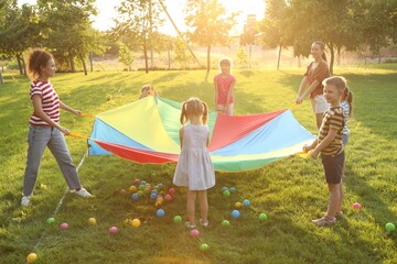 Naklejka na ściany i meble Group of children and teachers playing with rainbow playground parachute on green grass. Summer camp activity