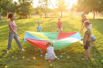 Group of children and teachers playing with rainbow playground parachute on green grass. Summer camp activity