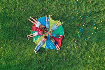 Group of children with teachers holding hands together on rainbow playground parachute in park, top...