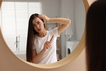 Emotional woman with comb examining her hair and scalp near mirror in bathroom. Dandruff problem