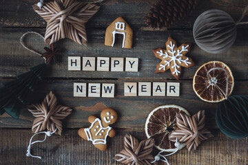 Holidays card on wooden background with gingerbread cookies, handmade craft toys, festive rustic zero waste decorations. Sustainable lifestyle. Happy New Year greeting written with wooden blocks.
