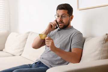 Emotional man with credit card talking on smartphone at home. Be careful - fraud