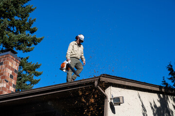 Fall pine needle debris flying in the air as a senior man on a rooftop is cleaning out gutters with...