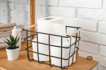 Basket with toilet paper rolls, plant and towels on wooden shelving unit near light brick wall, closeup