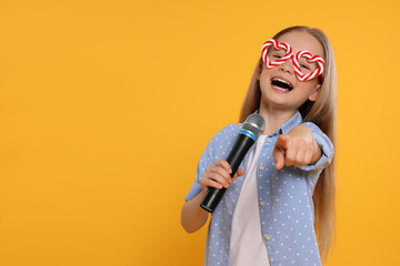 Cute little girl with funny glasses and microphone singing on yellow background, space for text