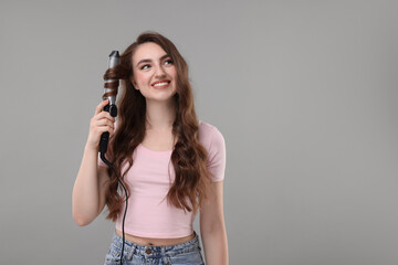 Beautiful young woman using curling hair iron on grey background, space for text