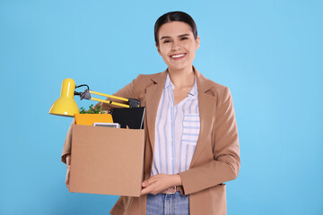 Happy unemployed woman with box of personal office belongings on light blue background