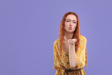 Beautiful young hippie woman blowing kiss on violet background, space for text