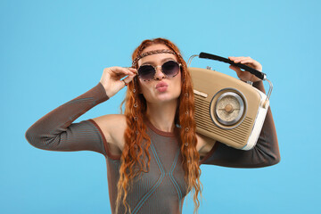 Stylish young hippie woman with retro radio receiver sending air kiss on light blue background