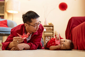 Portrait of young Asian man as father playing with cute baby boy lying on floor at home and holding...