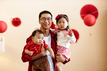Waist up portrait of happy Chinese father holding two children and smiling at camera with red paper...