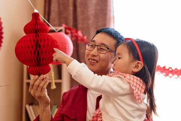 Portrait of cute Asian little girl hanging red paper lanterns at home with father preparing for...