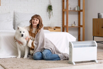 Young woman with plaid and Samoyed dog warming near radiator in bedroom