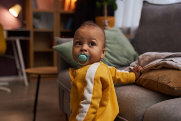 Portrait of cute baby boy with binky taking first steps in cozy living room, copy space
