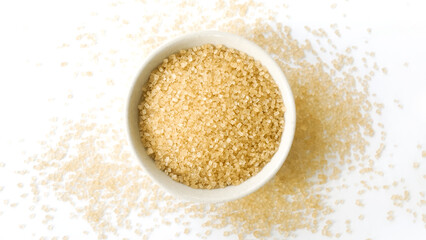 Small bowl of raw brown sugar with scattered crystals on white background