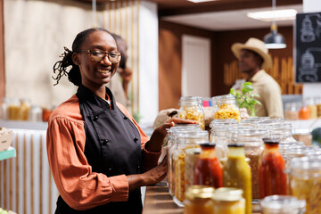 Smiling woman standing next to a wooden shelf filled with bulk food glass containers. These...