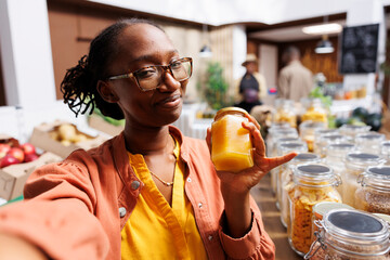 During video call, african american lady with glasses clutches a glass jar of honey. At eco...