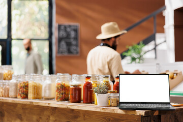Digital laptop with blank white screen is placed next to organic bulk products in jars on shelves. On the wooden counter is a minicomputer running an isolated display with a mockup copyspace template.