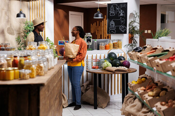 African American shopkeeper and customer discuss eco friendly, pantry staples and fresh produce. Sustainable shopping, bulk items, and plastic-free standards. Focus on background. - obrazy, fototapety, plakaty