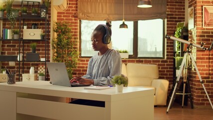 Fototapeta na wymiar Freelancer enjoys working at workstation while listening to music and performing internet activities. At sunset, african american woman is multitasking, online career in blogging.