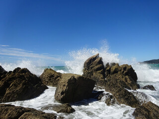 Fototapeta na wymiar Tosende Welle schlägt gegen Felsen in der Brandung im Meer mit blauem Himmel im Hintergrund