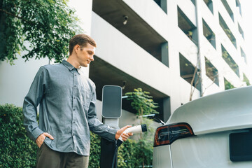 Young man travel with EV electric car charging in green sustainable city outdoor garden in summer...