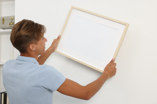 Young man hanging picture frame on white wall indoors, back view