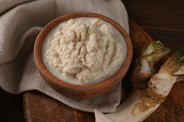 Spicy horseradish sauce in bowl and root on wooden table, closeup