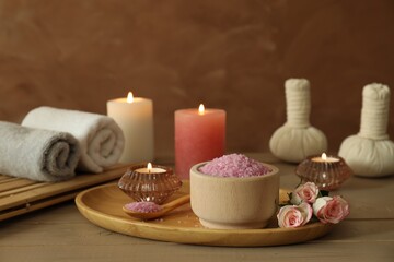 Fototapeta na wymiar Bowl of pink sea salt, roses, burning candles, herbal massage bags and towels on wooden table