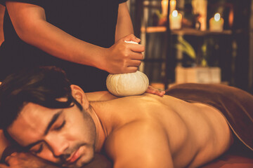 Hot herbal ball spa massage body treatment, masseur gently compresses herb bag on man body. Tranquil and serenity of aromatherapy recreation in warm lighting of candles at spa salon. Quiescent - obrazy, fototapety, plakaty