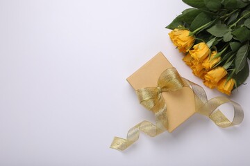 Gift box with golden bow and bouquet of beautiful roses on white background, flat lay. Space for text