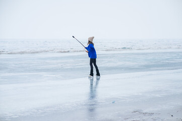 A caucasian woman is skating on a frozen lake holding a selfie stick in her hands. The figure skater films her skating.