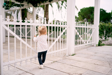 Little girl stands holding on to the bars of a metal fence in the park and looks into the distance. Back view