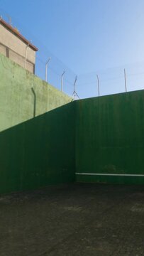 Explore the dynamic ambiance of a concrete-floored court wall, camera gliding towards the towering dark green walls adorned with a low white horizontal line. Shadowed elegance under a clear, blue sky.
