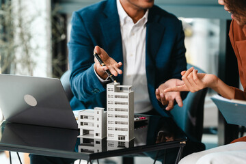 Attractive Asian businessman and African American female real estate and land investor talk discussing together on table with laptop and small building model. Reserve fund. - obrazy, fototapety, plakaty