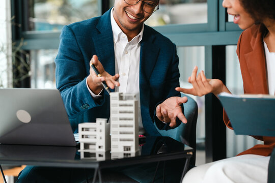 Attractive Asian businessman and African American female real estate and land investor talk discussing together on table with laptop and small building model. Reserve fund.