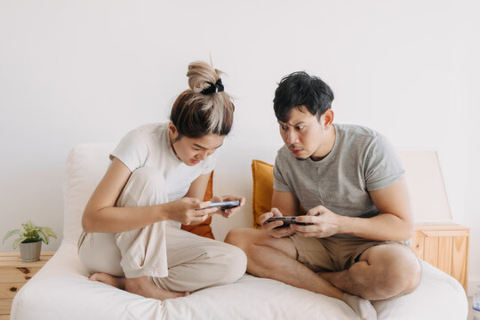 Asian Thai Couple Using Mobile Phone Together White Sitting On White Sofa In Apartment Room, Playing Game Online.